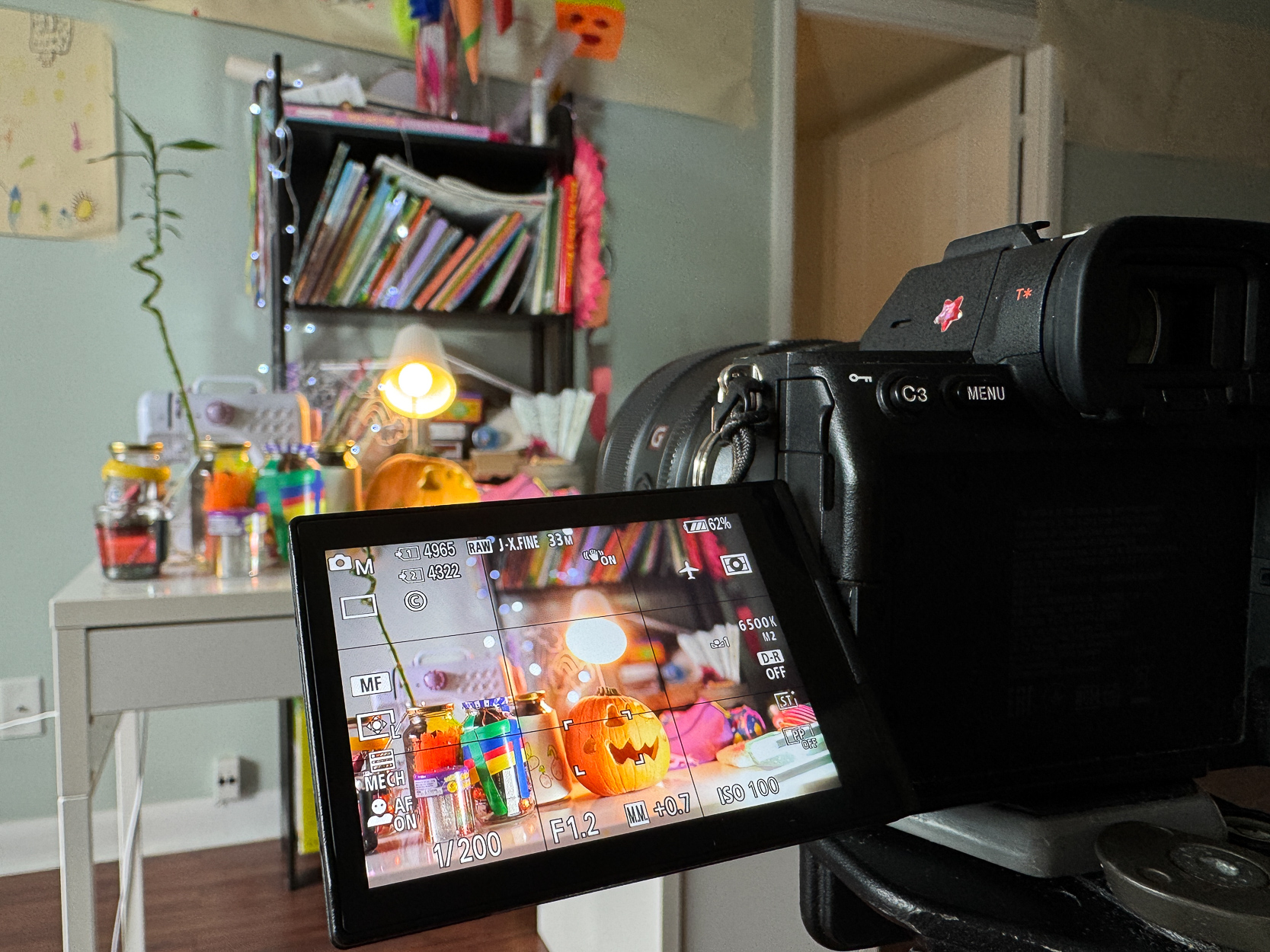 A camera captures a vibrant desk setup with colorful jars, books, and a lamp, viewed on its display screen. The scene includes Halloween decor with a pumpkin, highlighting a cozy and creative workspace.