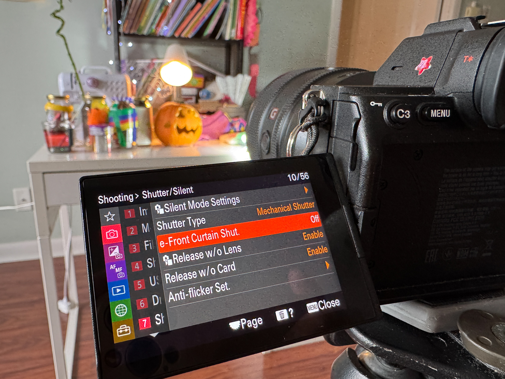 A digital camera displays its settings menu on the monitor, featuring options like silent mode and shutter type, with a cozy Halloween-themed background including a carved pumpkin and colorful jars on a desk.