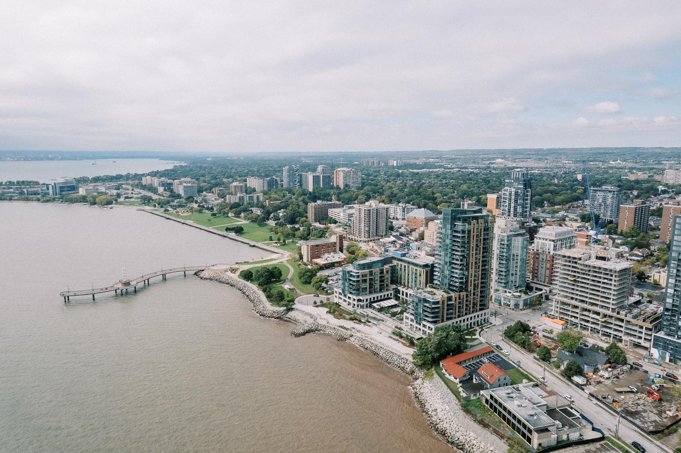 Aerial view of an urban waterfront area with modern high-rise buildings and a scenic boardwalk along the water, set against a backdrop of lush greenery and a cloudy sky.
