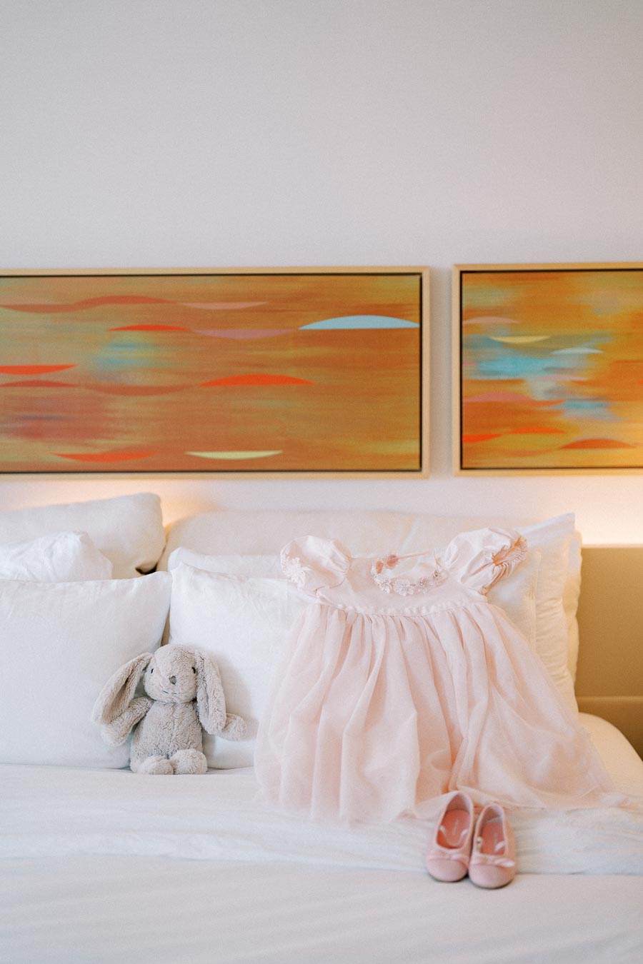 Child's pink dress with floral details and matching shoes on a white bed, next to a plush toy bunny. Two abstract paintings with warm colors hang above the bed.