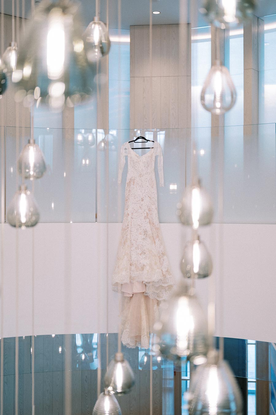 Elegant lace wedding dress hanging in a modern room with soft lighting and decorative glass pendants in the foreground.