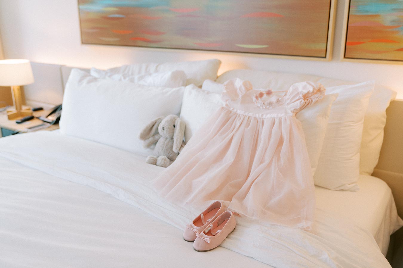 A soft pink baby dress and matching shoes elegantly placed on a white bed next to a plush bunny toy, creating a serene and cozy nursery setting.