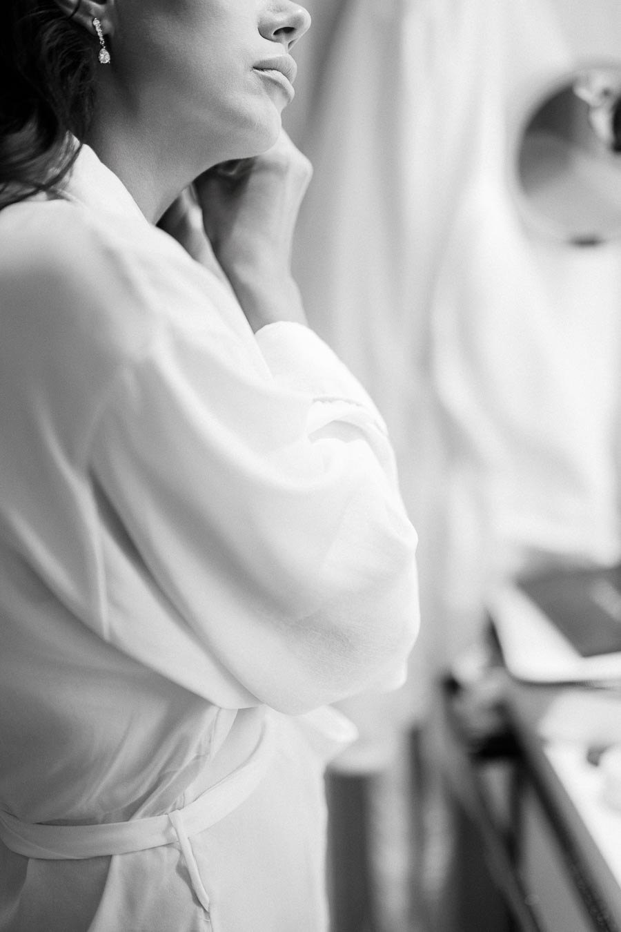 A black and white image of a woman in a bathrobe putting on an earring in front of a mirror, capturing a serene and elegant moment of self-preparation.