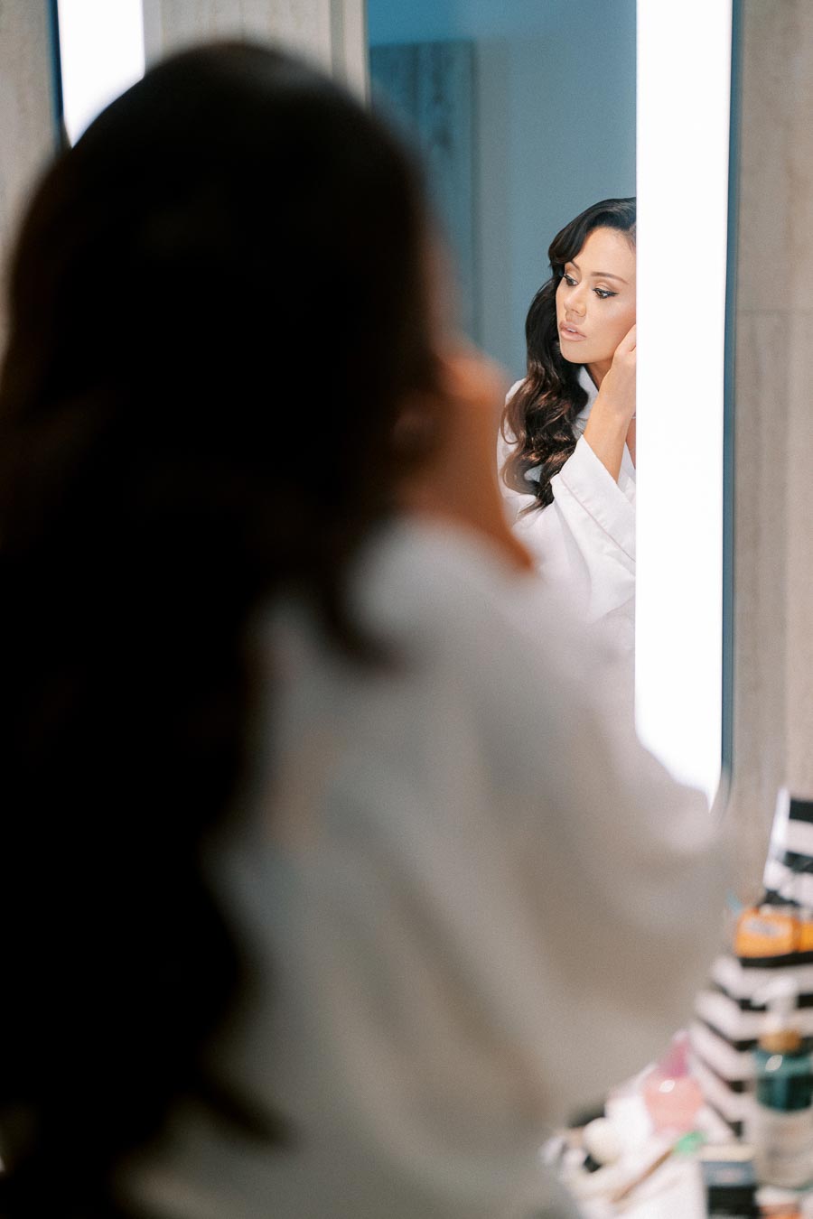 Elegant woman in white robe styles hair in front of illuminated bathroom mirror, surrounded by skincare products.