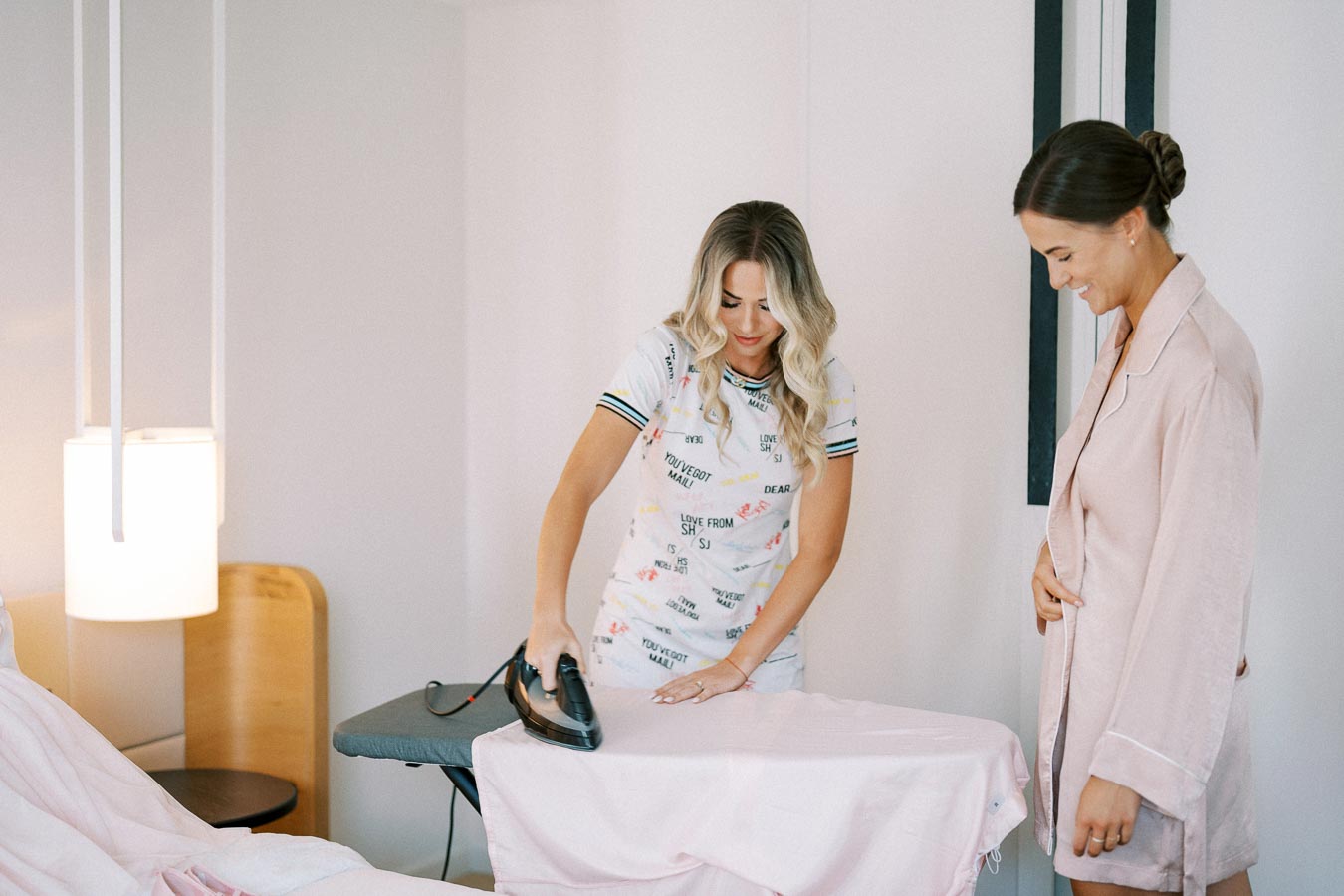 Two women in a bedroom setting with one ironing a shirt while the other watches, highlighting home tasks and domestic lifestyle.