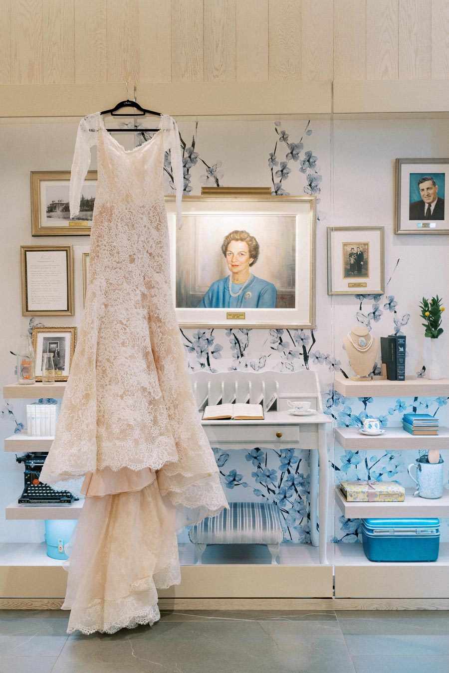 A lace wedding dress hanging in a beautifully decorated room with framed portraits, floral wallpaper, and various artifacts on shelves, creating an elegant and nostalgic atmosphere.