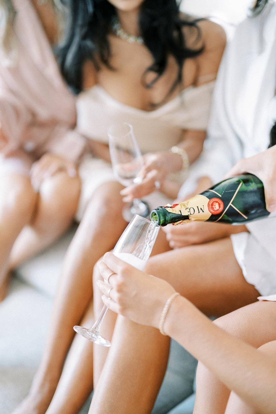 Women celebrating with champagne, pouring Moët & Chandon into a flute at a bachelorette party, wearing elegant dresses and jewelry.