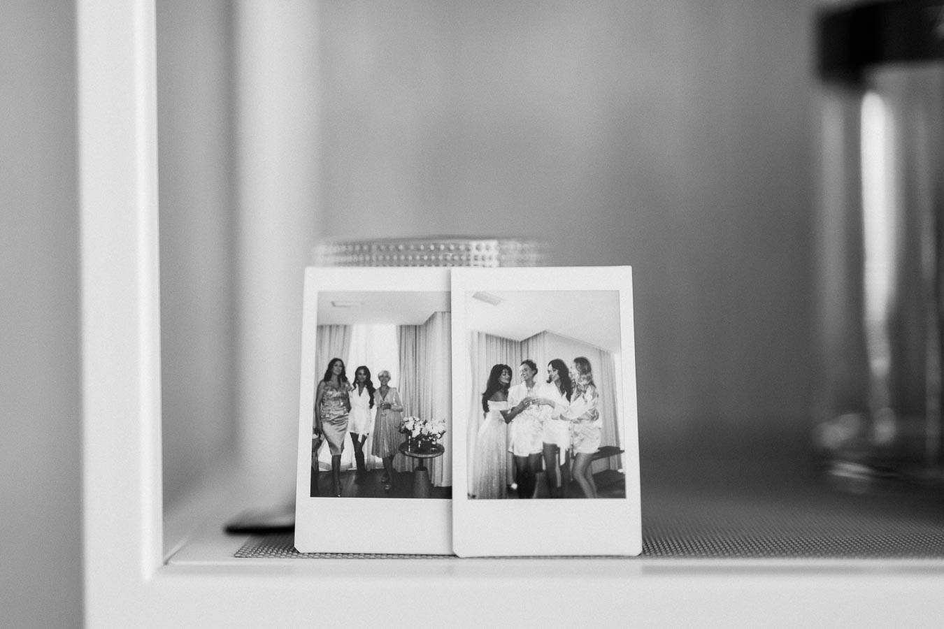 Black and white photo of two polaroid images displayed on a shelf, featuring a group of women smiling and celebrating together indoors.