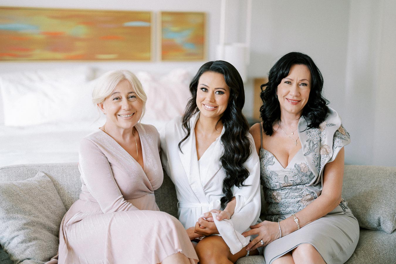 Three women smiling and sitting on a sofa, wearing elegant dresses in a bright, softly lit room with abstract artwork in the background. Perfect for lifestyle or family-themed content.