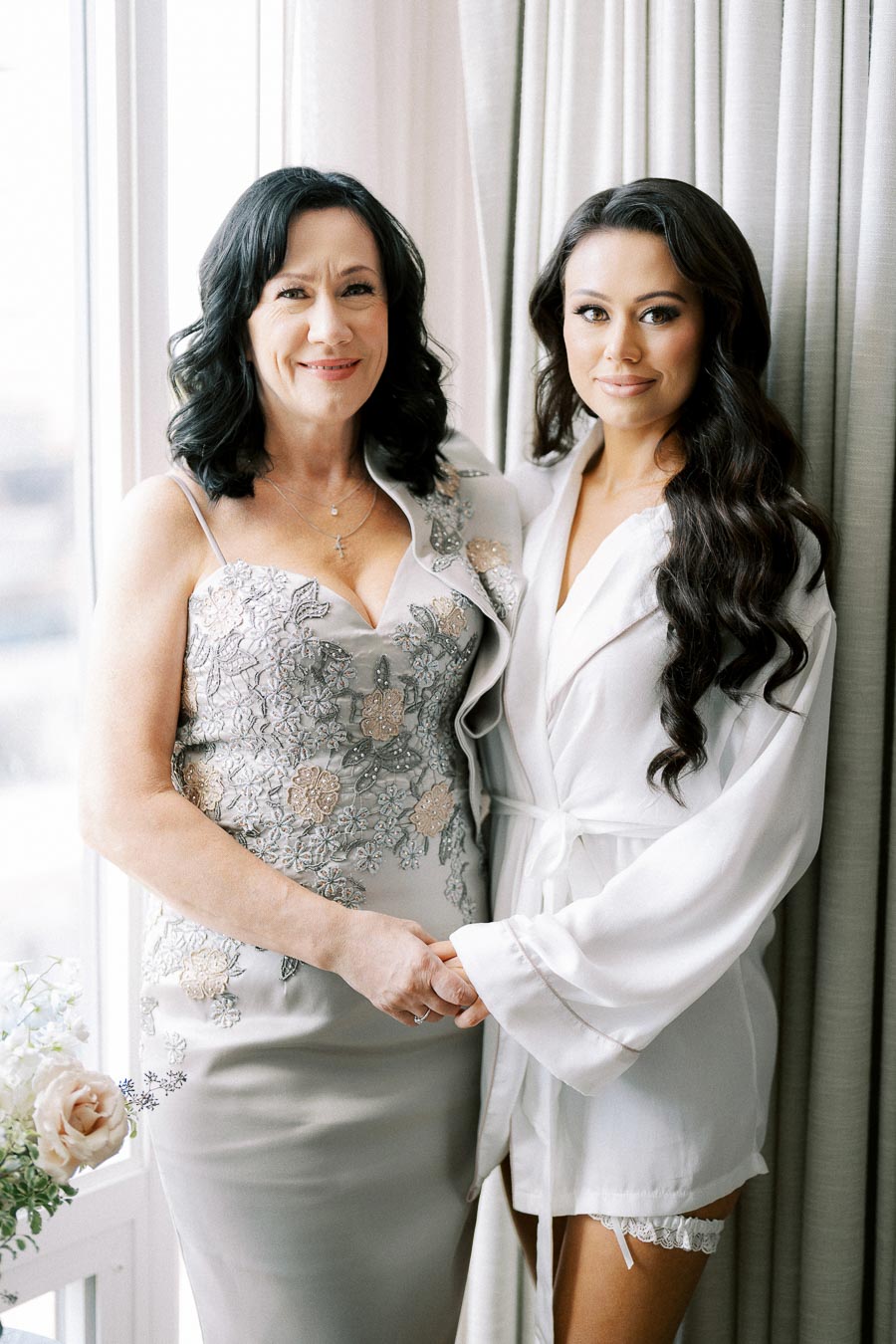 A mother and daughter share a tender moment before a wedding, with the mother wearing a floral-embroidered dress and the daughter in a bridal robe, standing by a sunlit window with flowers in the foreground.