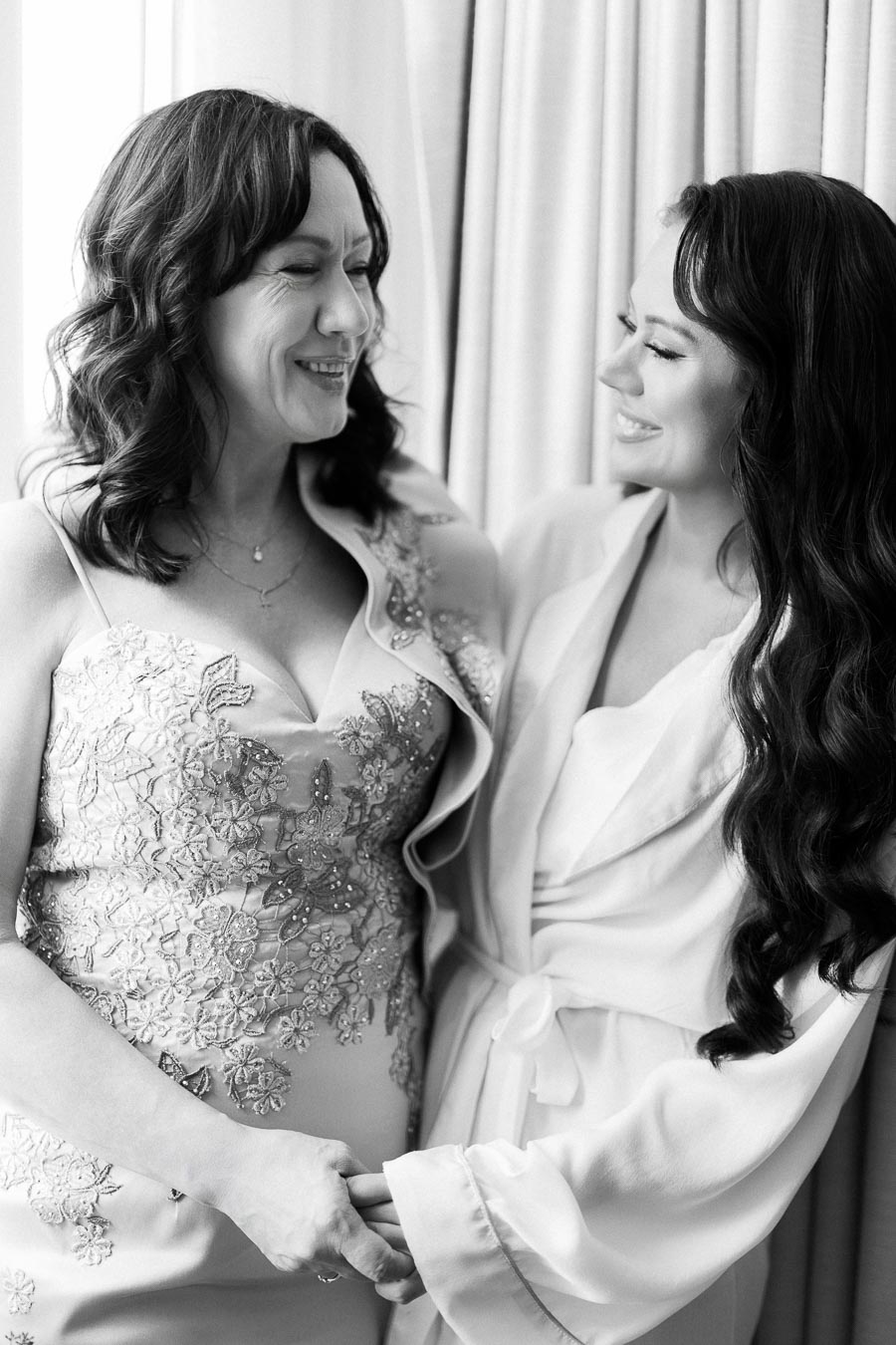 A bride and her mother share a joyful moment, holding hands and smiling at each other while preparing for a wedding day. The bride wears a robe, and her mother wears an embroidered dress, conveying warmth and love. Black and white photo.