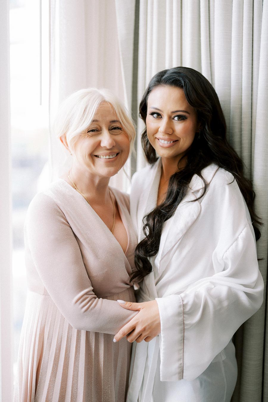 A bride in a white robe stands beside an older woman in a light pink dress, both smiling warmly near a window with soft curtains.