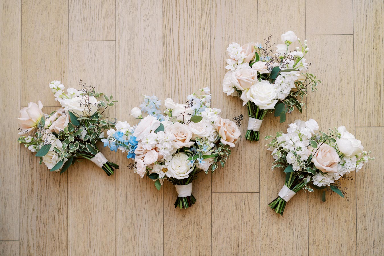 Elegant floral bouquets featuring blush pink and white roses with delicate greenery, arranged on a light wood floor. Perfect for weddings or special occasions.