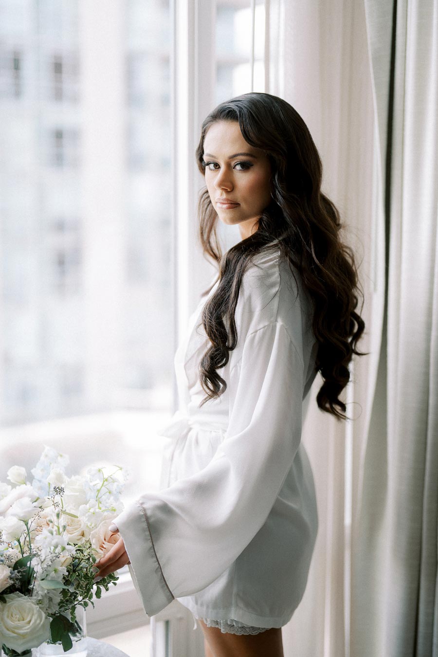 A woman in a white silk robe stands by a window, gently touching a bouquet of white roses and greenery, with soft daylight illuminating her long, dark wavy hair and serene expression.