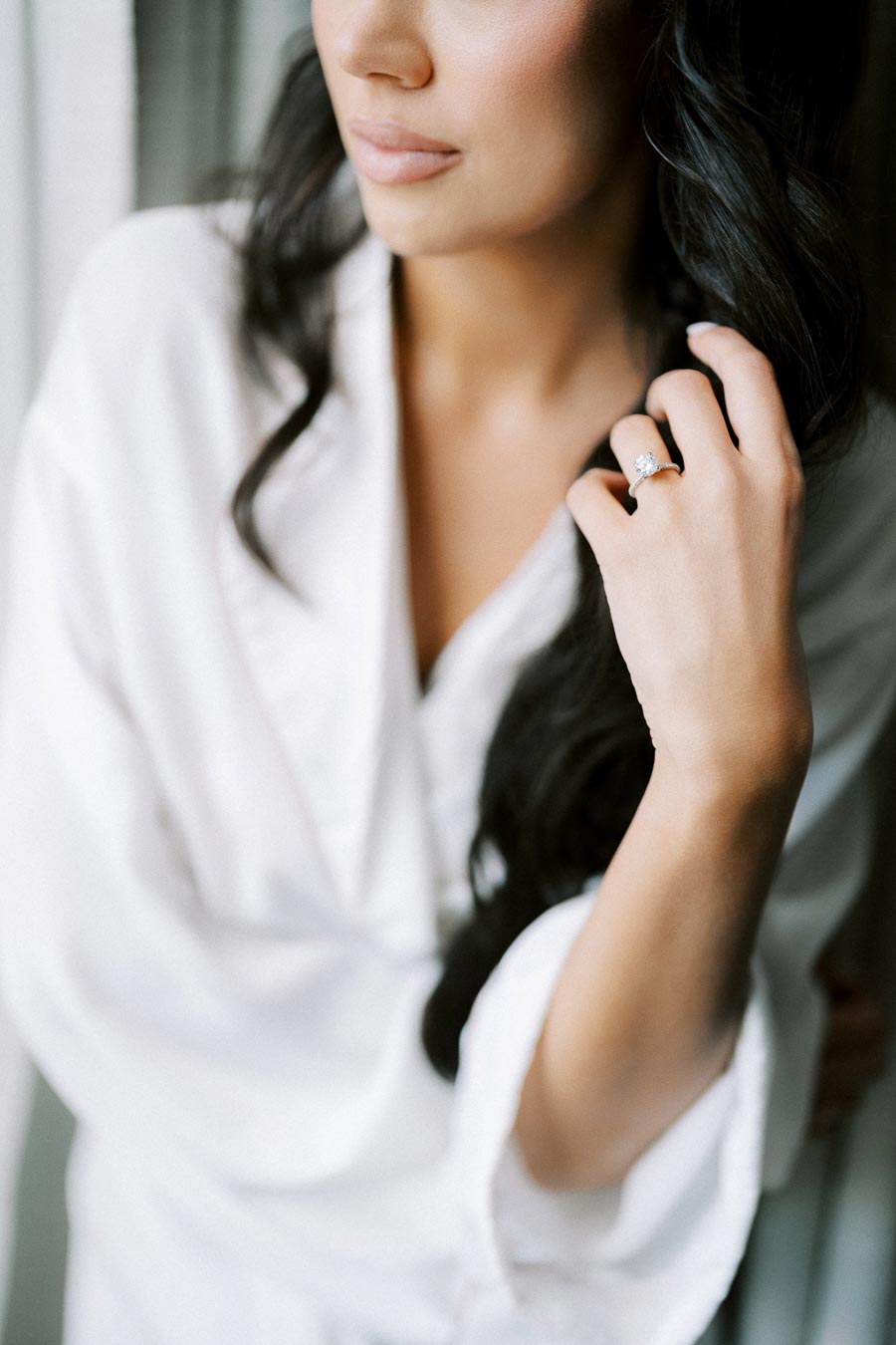 Woman wearing elegant white robe admiring her engagement ring, showcasing a minimalist and bridal style.