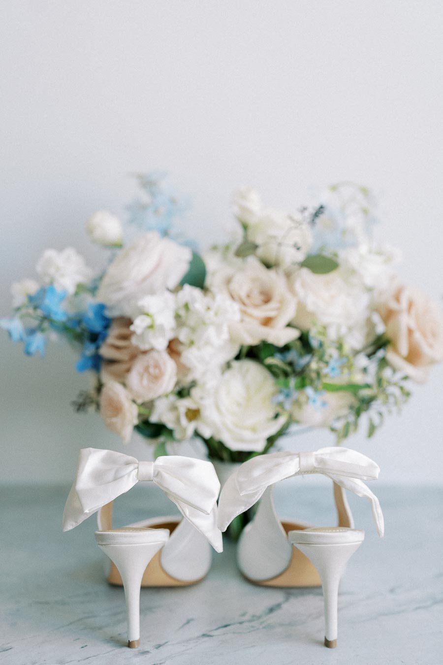 Elegant white high heels with bows next to a pastel floral bouquet on a marble surface, perfect for a chic wedding theme.