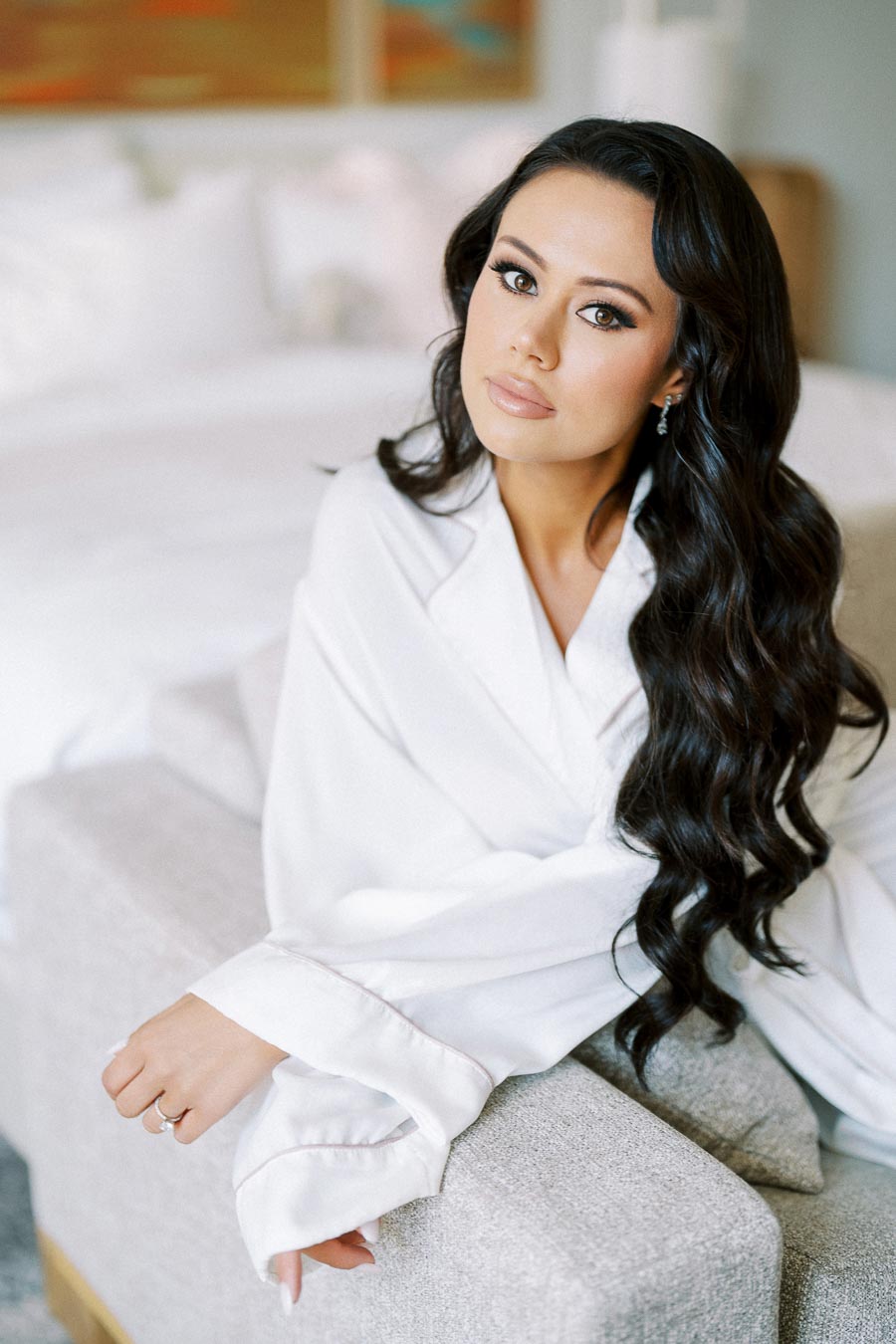 Elegant woman in white robe sitting on sofa with long flowing dark hair, soft bedroom background.