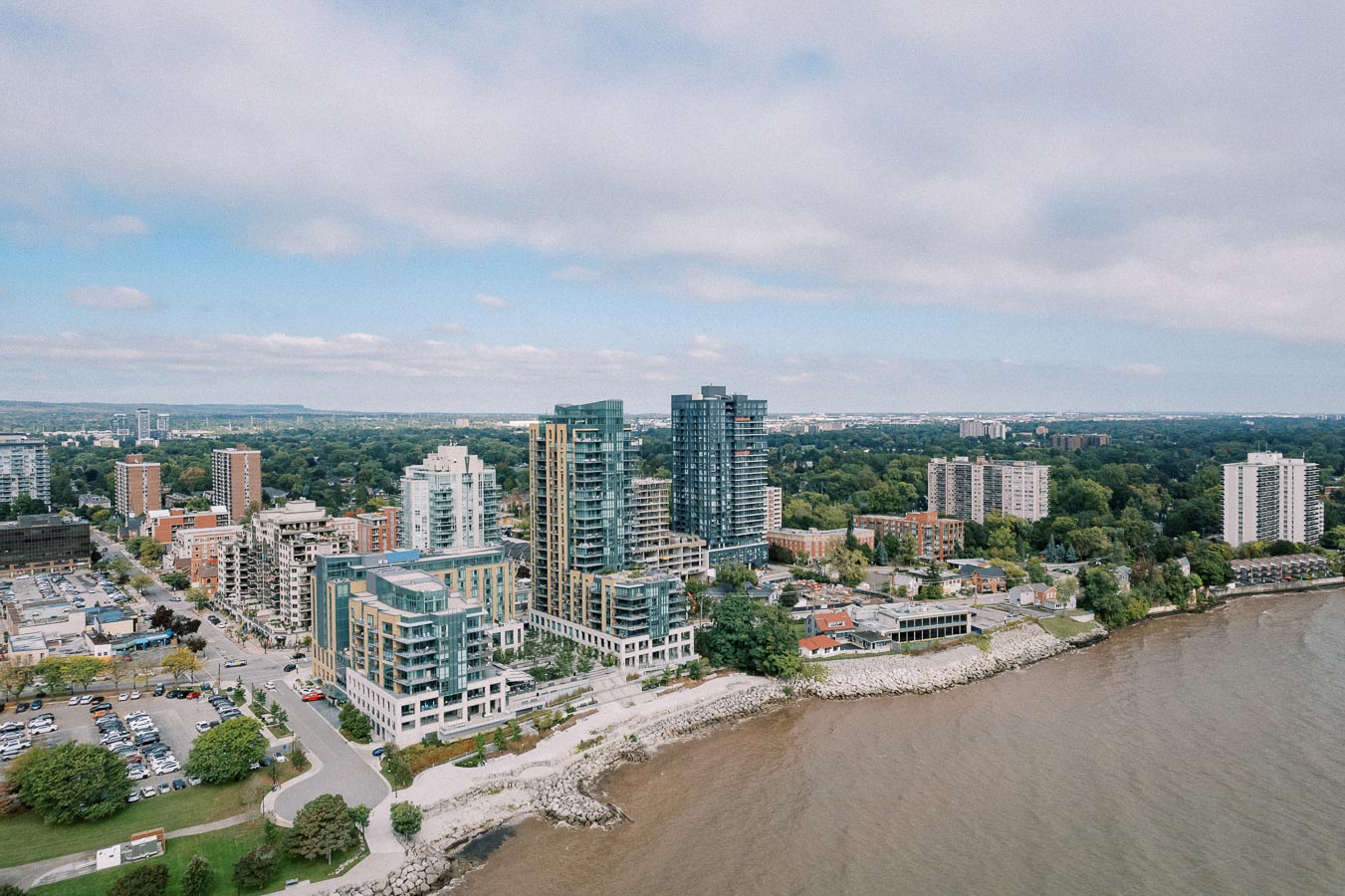 Aerial view of a waterfront cityscape featuring modern high-rise buildings and lush greenery, under a partly cloudy sky, with a body of water in the foreground.