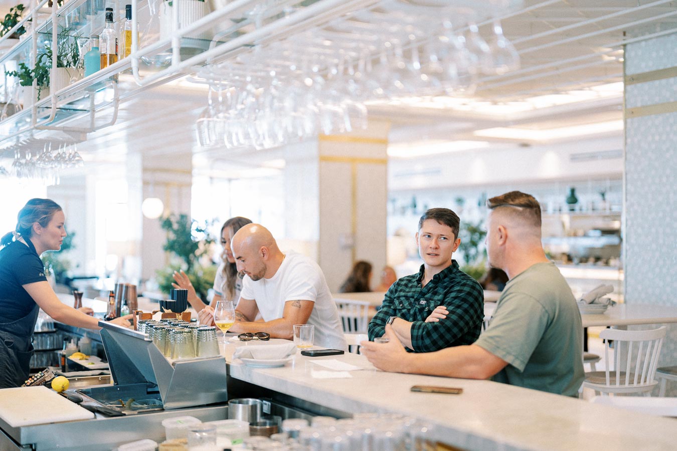 People sitting at a modern bar during the day, engaging in conversation while a bartender serves drinks, in a brightly lit and stylish cafe setting.