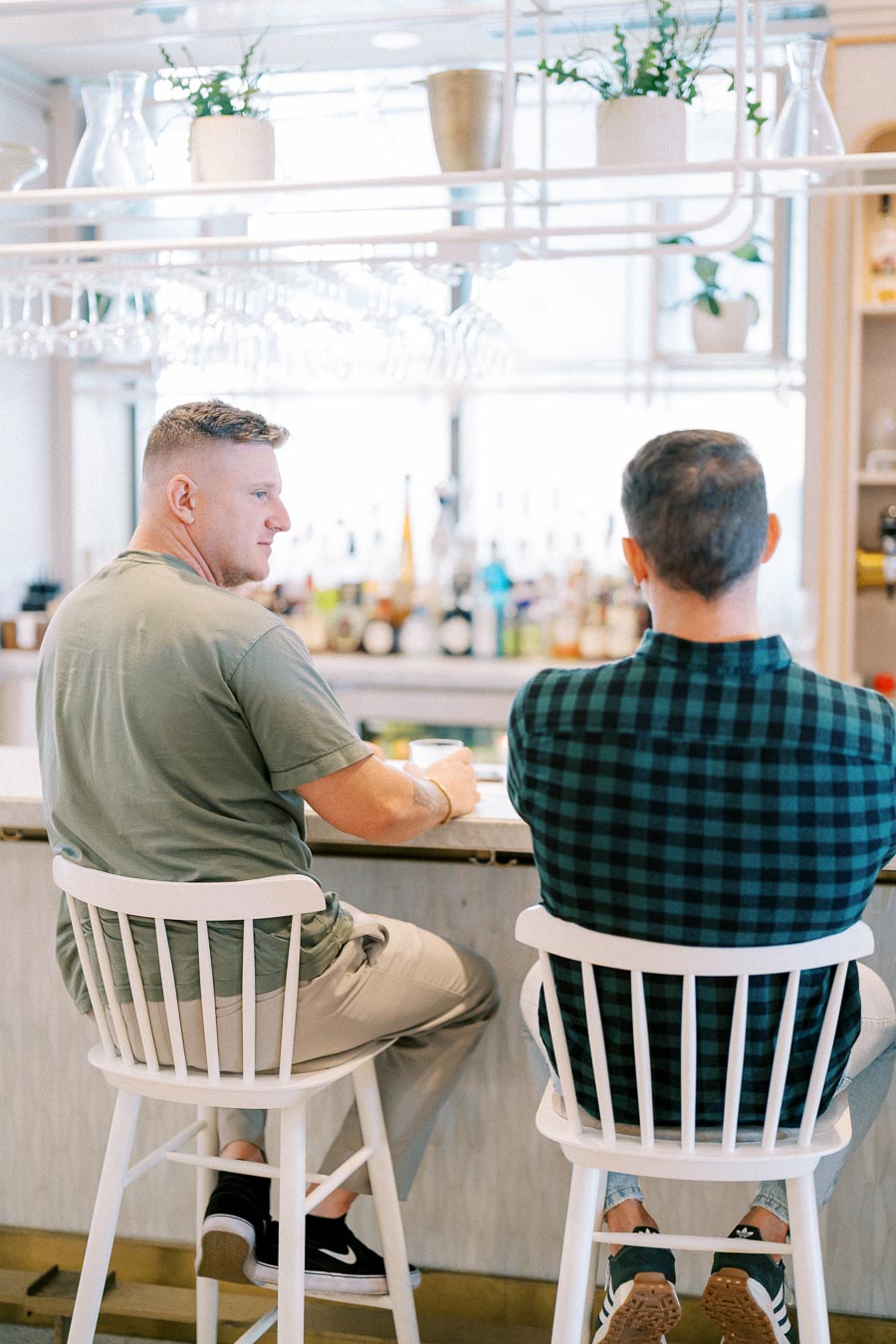 Two men sitting on white bar stools at a bright, modern bar with glassware and potted plants overhead, engaging in conversation.