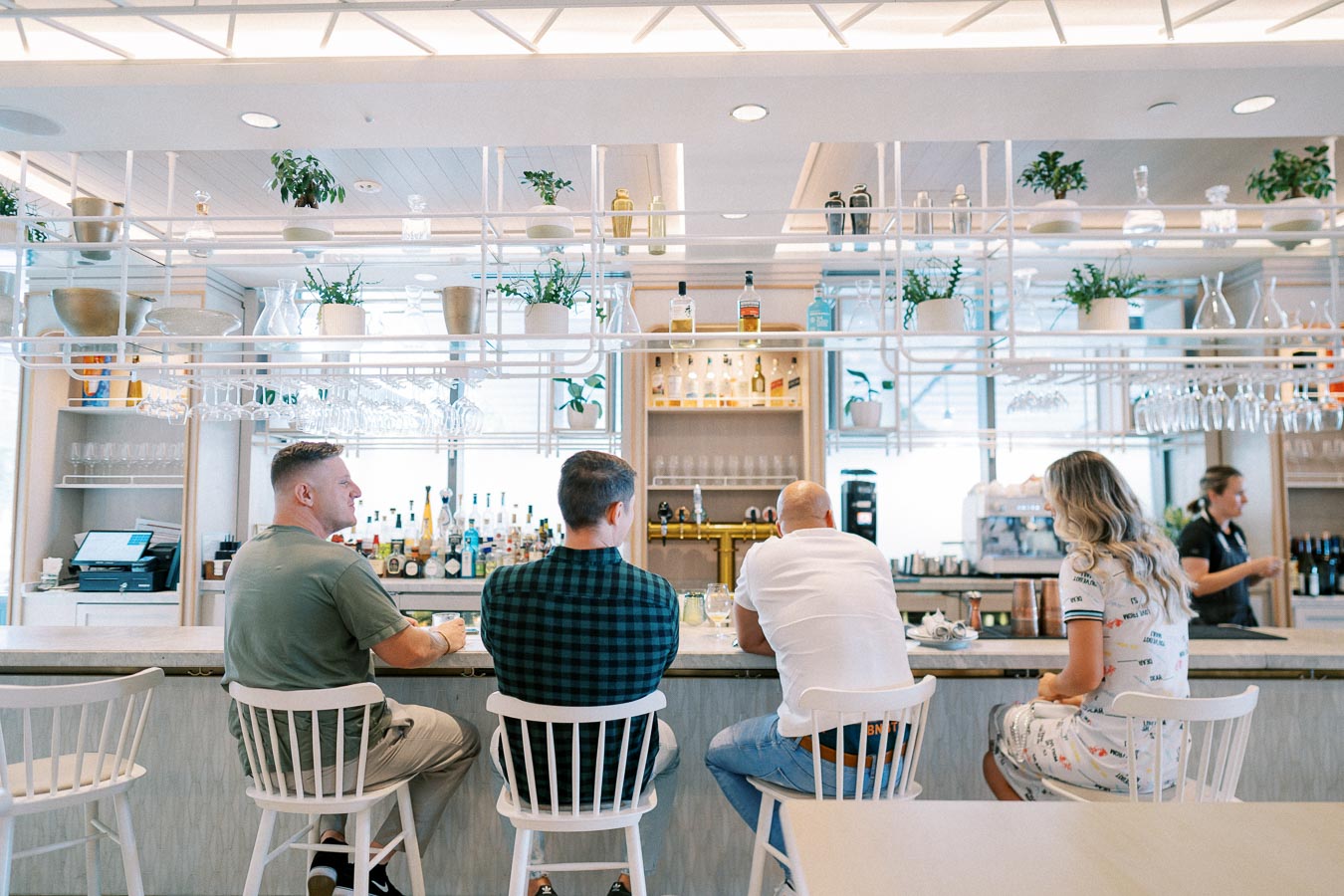 Modern cafe interior with patrons sitting at a stylish bar counter, surrounded by hanging plants and a variety of bottles, creating a welcoming and trendy atmosphere.