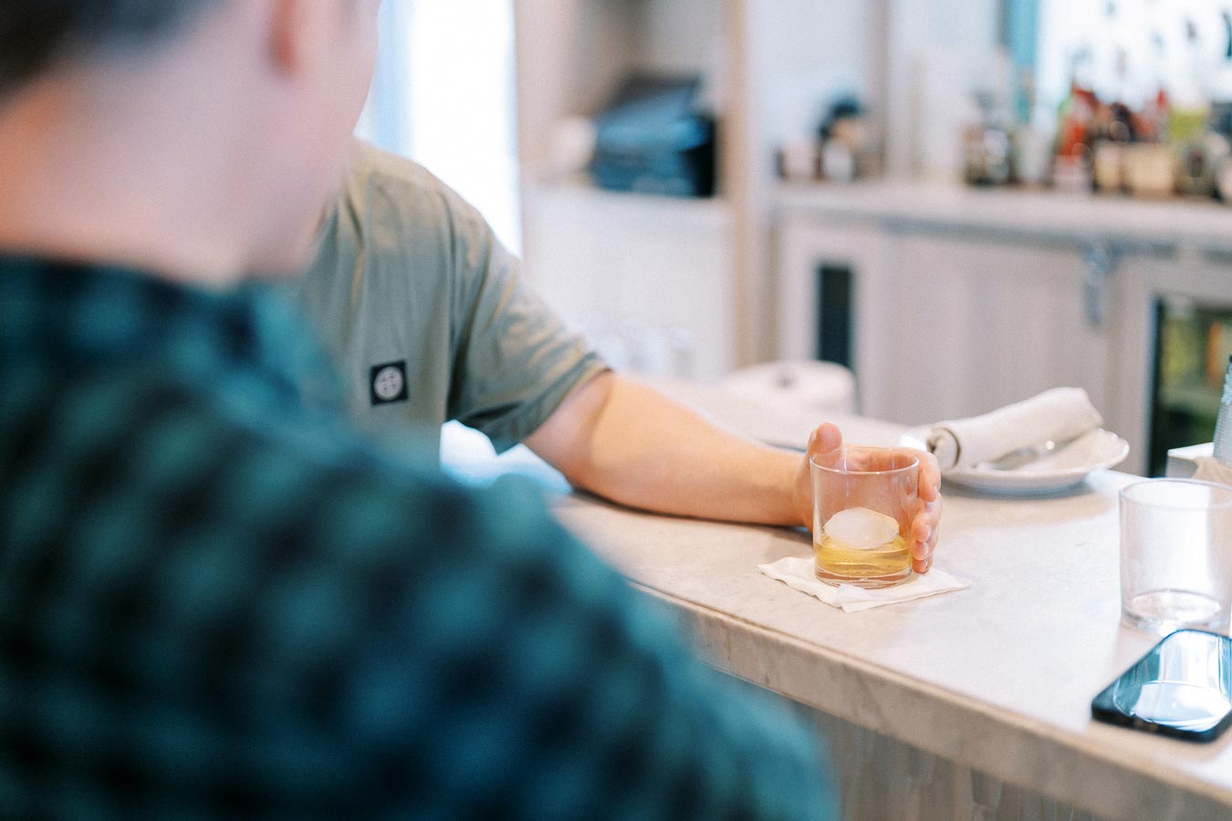 A person holding a glass of whiskey with ice at a bar counter, with a blurred background of bar items and decor.