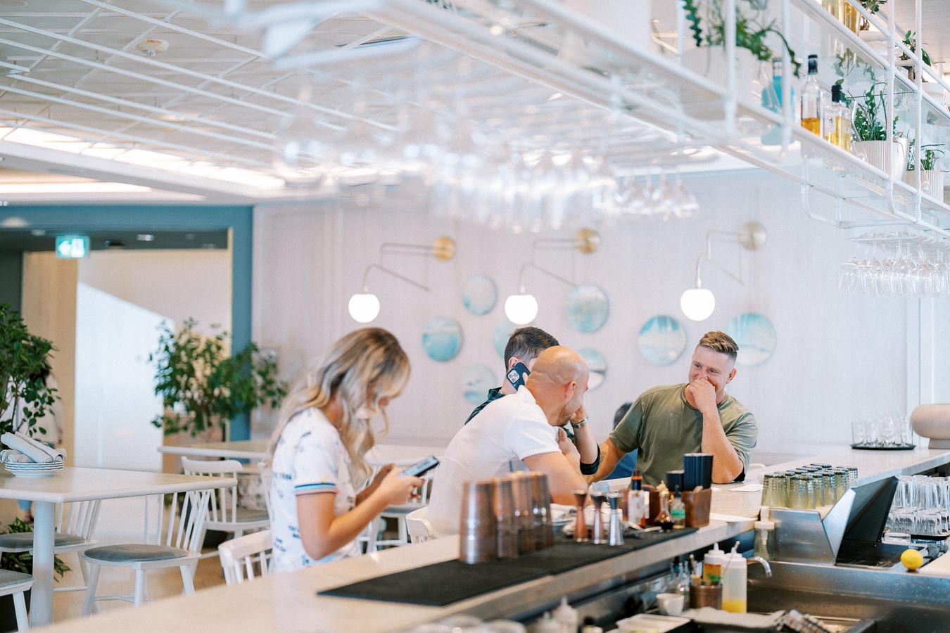 Modern cafe interior with people socializing at the bar, featuring stylish decor, potted plants, and hanging glasses.