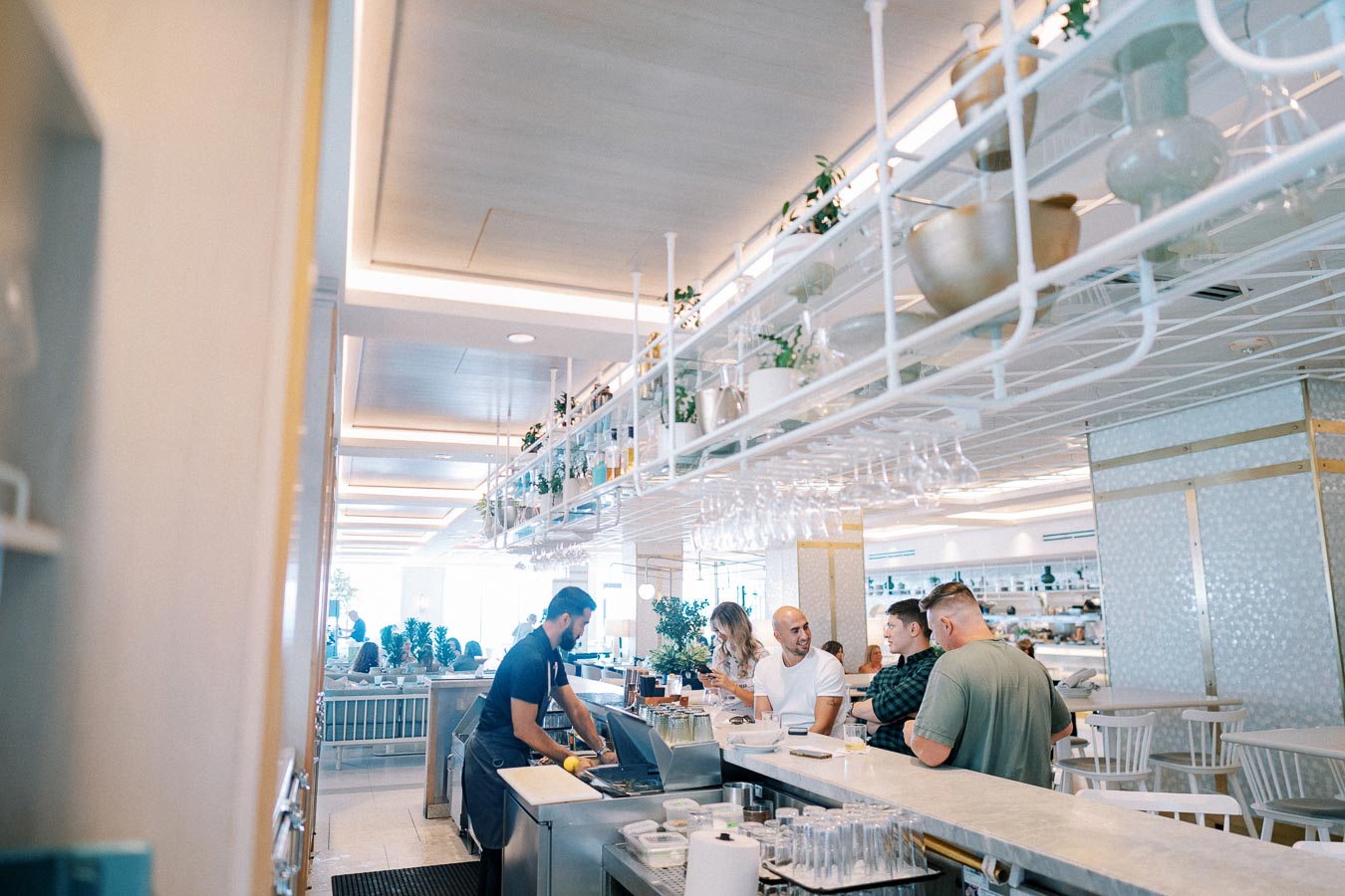Modern restaurant interior with patrons at a stylish bar counter, where a bartender wearing an apron prepares drinks, surrounded by hanging plants and glassware.