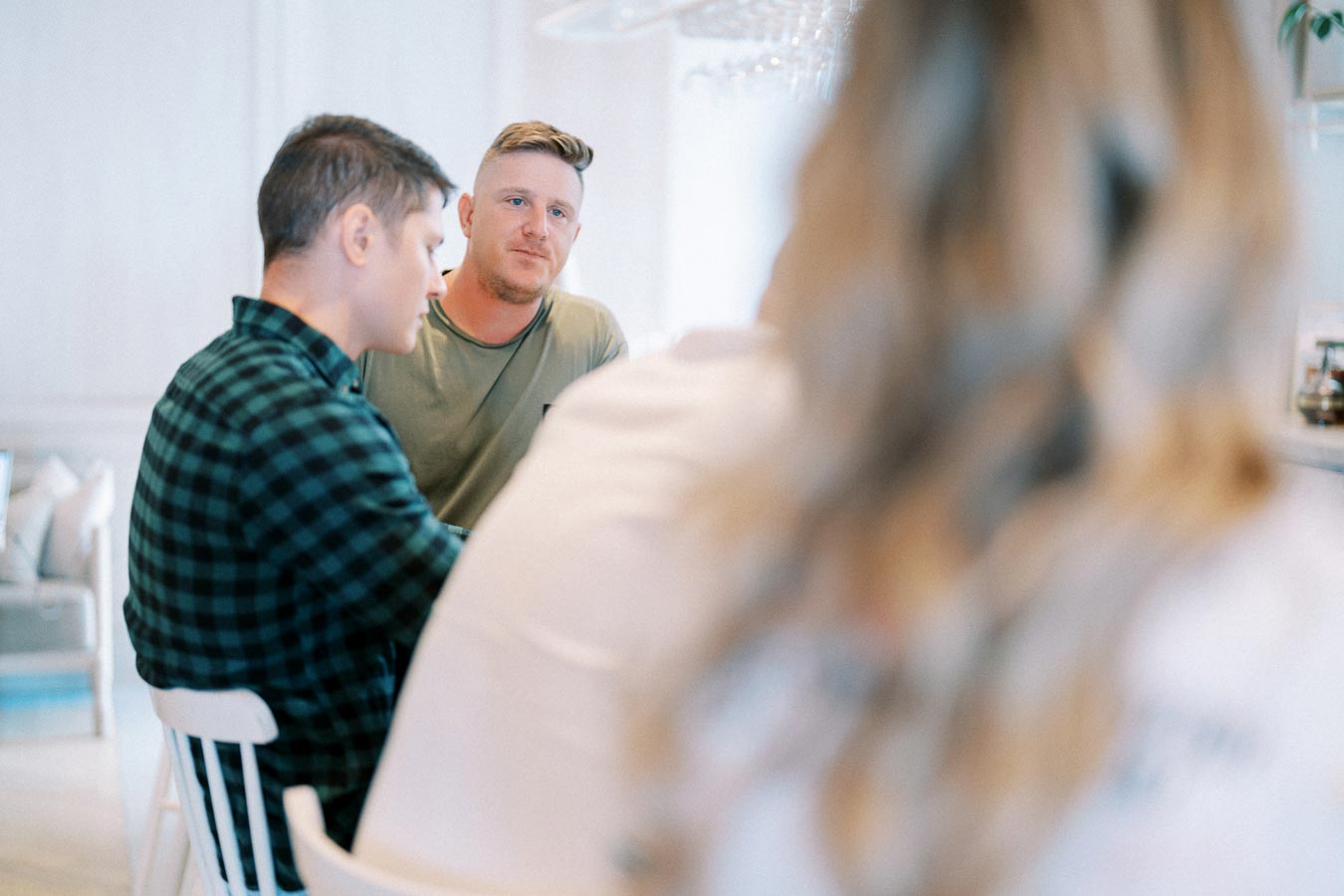 A group of people engaged in a business discussion around a table, two men in focus, with blurred foreground. Bright and modern office setting, capturing collaborative teamwork.