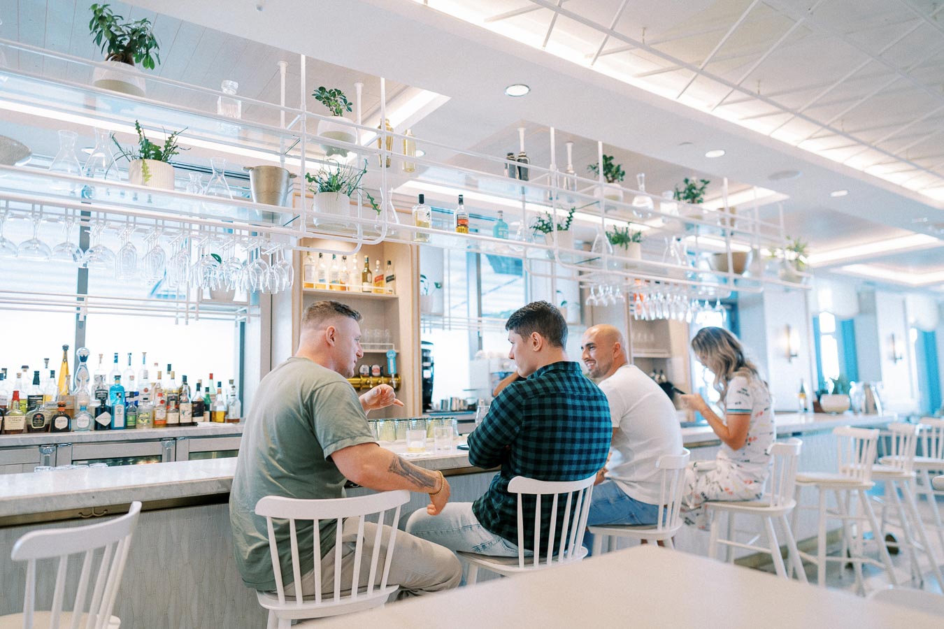Alt text: Group of people enjoying drinks and conversation at a modern bar with a stylish interior, featuring white chairs and hanging plants.
