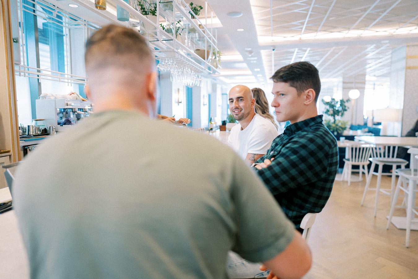 Three men sitting at a modern cafe bar, engaging in conversation while surrounded by contemporary decor and natural lighting.