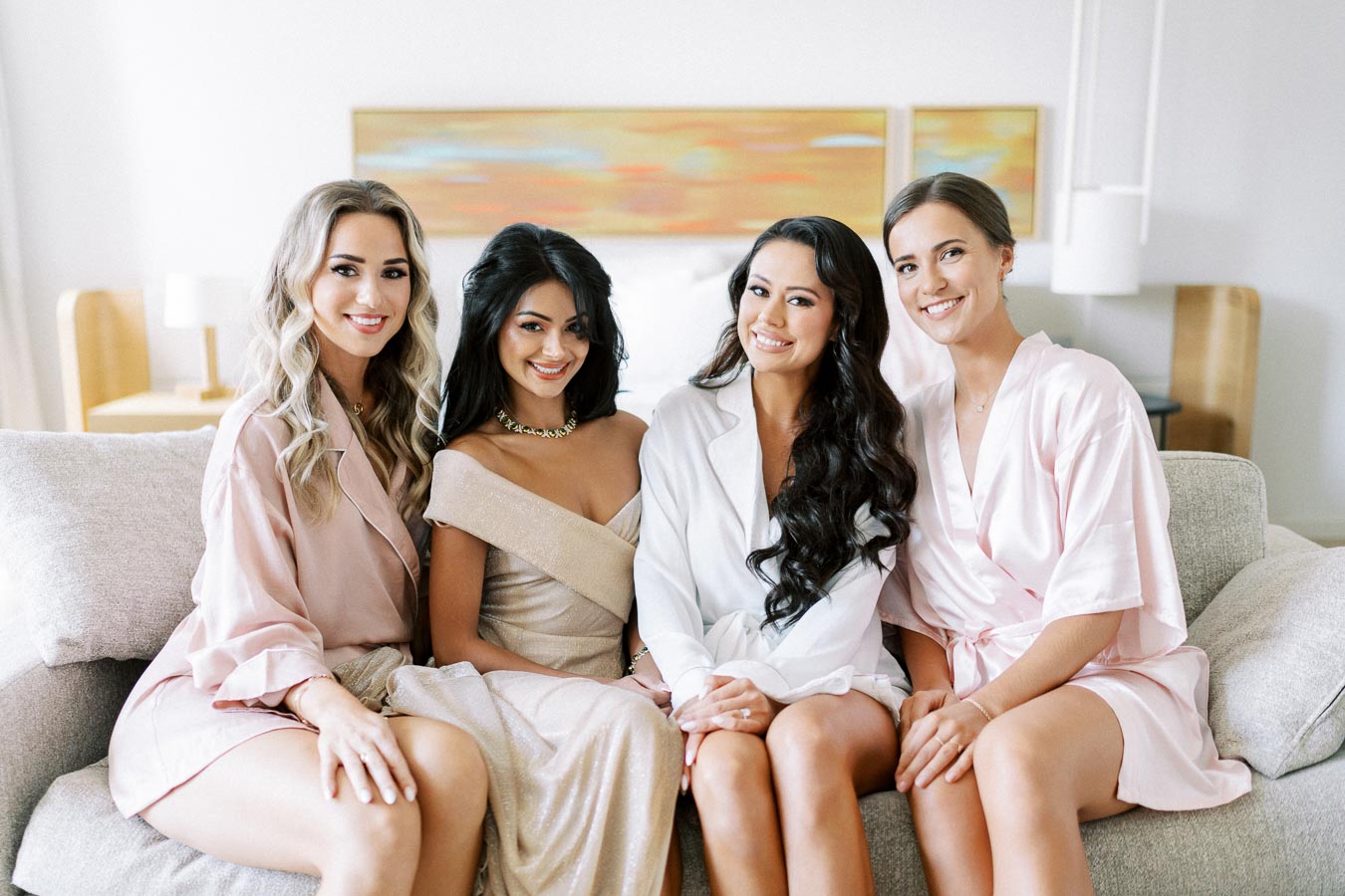 Four women smiling and sitting together on a couch, dressed in elegant robes, creating a warm and joyful atmosphere, perfect for a bridal party or women's gathering.