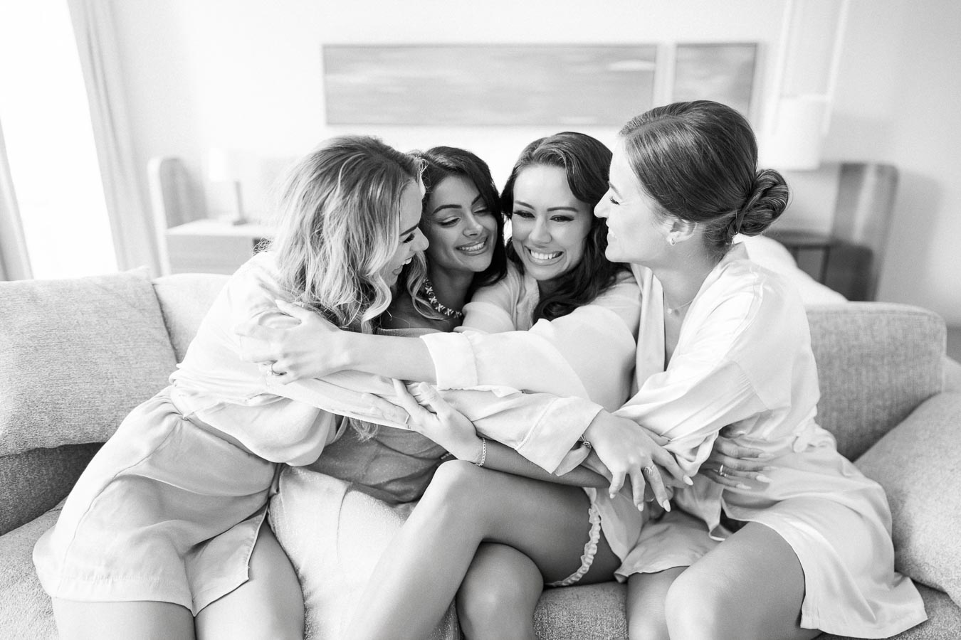 Four women in silk robes sharing a joyful group hug on a sofa, expressing friendship and happiness in a bright, cozy living room.