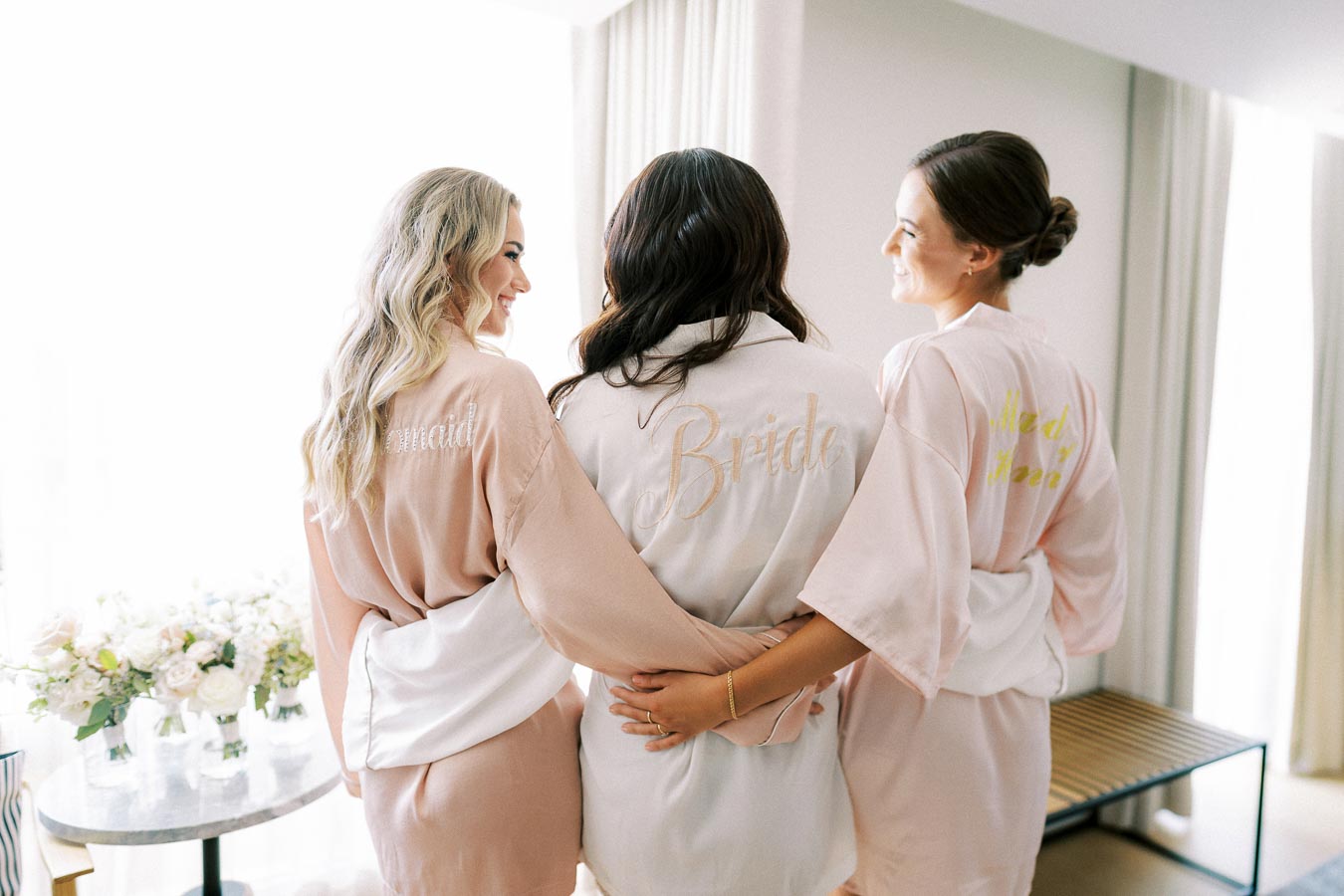 Bride and bridesmaids in personalized robes embrace on a wedding morning, surrounded by sunlight and floral arrangements.