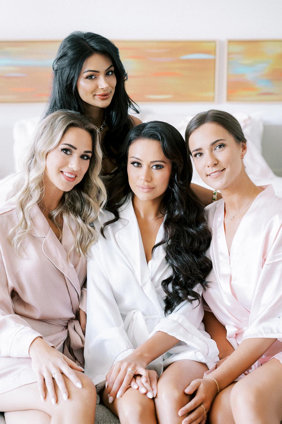 Four women dressed in pastel robes sitting together, smiling in a cozy indoor setting, perfect for a bridal party or getting ready scene.
