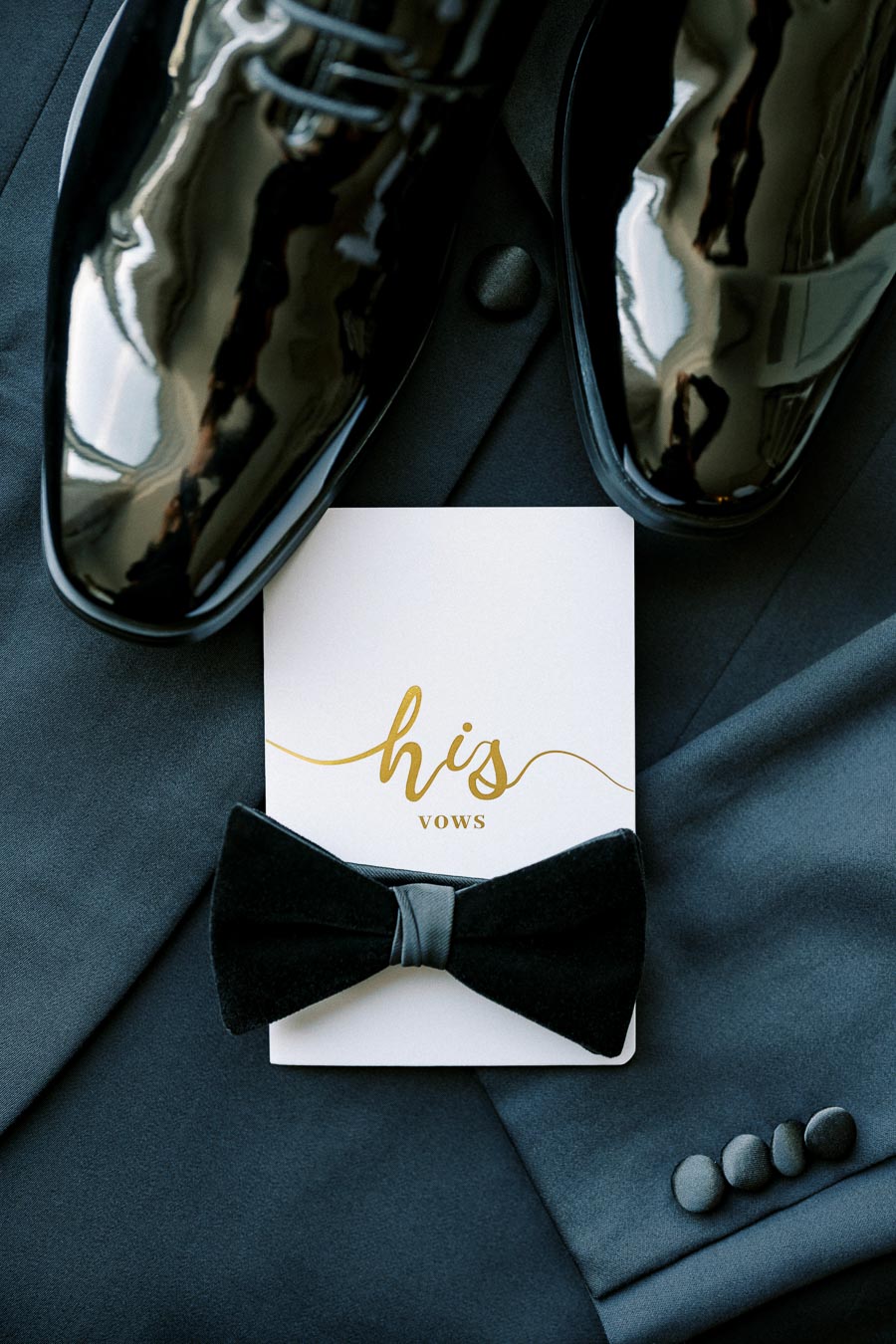 Black leather wedding shoes, a black bow tie, and a card labeled his vows elegantly arranged on a dark suit jacket, perfect for a groom's wedding preparation scene.