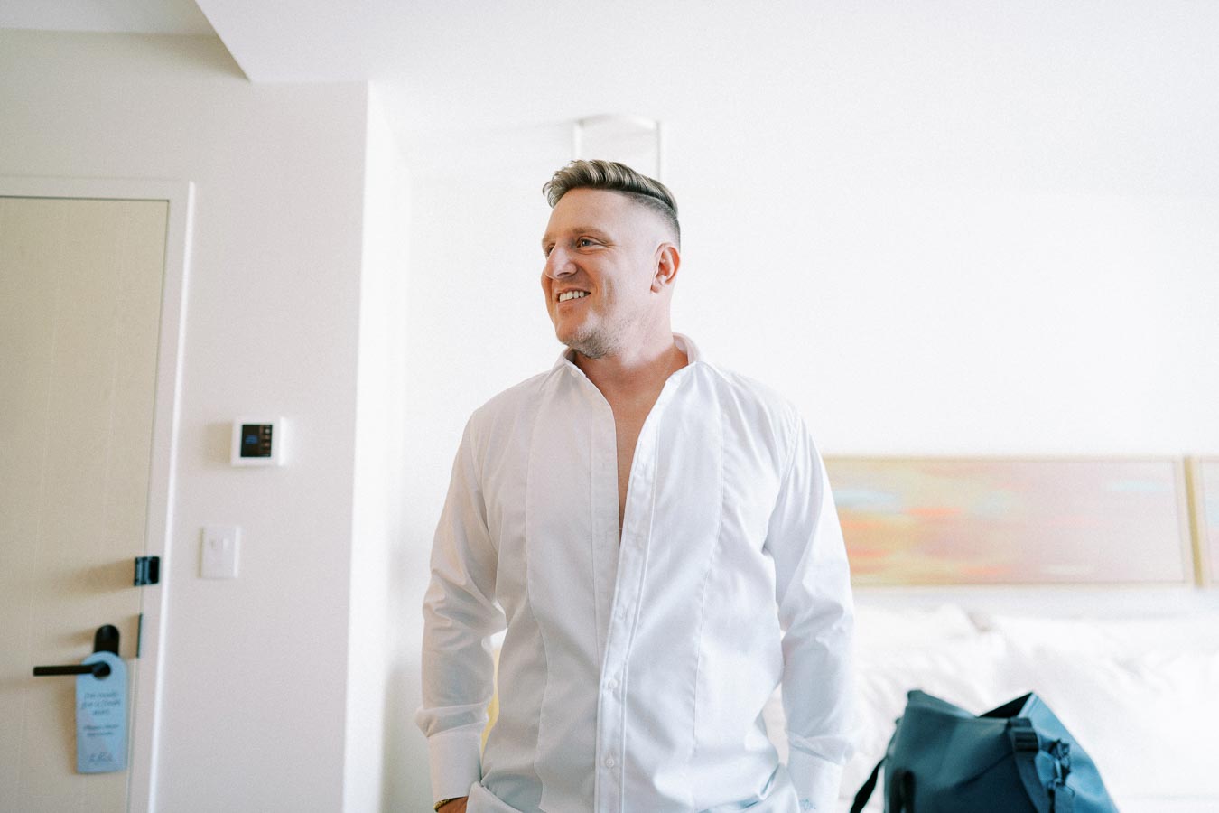 A man in a white shirt smiling in a bright hotel room with a suitcase nearby.