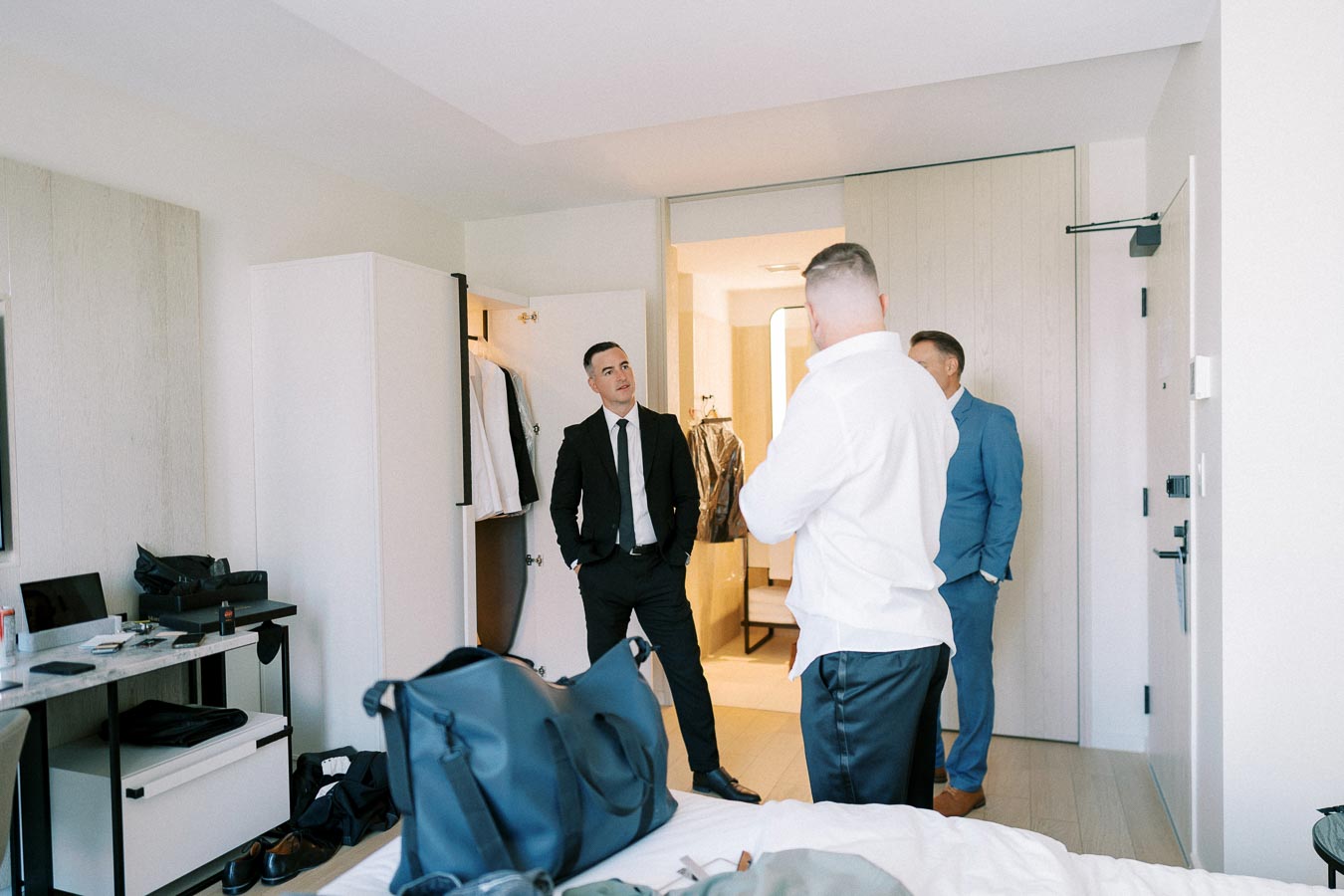 Men preparing for a formal event in a hotel room, with one in a suit and others getting ready. Suitcases and shoes are visible on the bed and floor, highlighting travel and preparation for a special occasion.