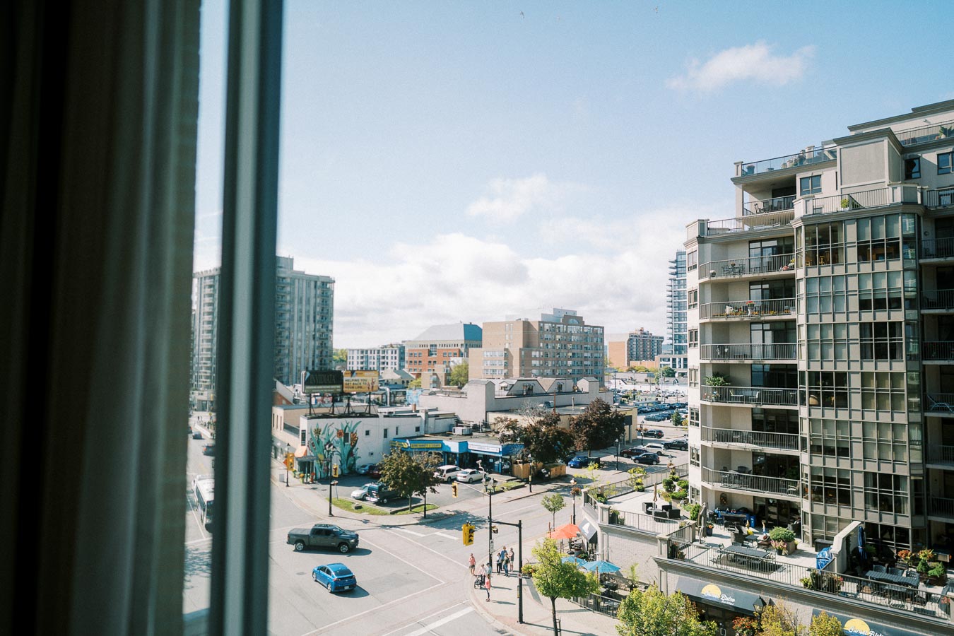 A vibrant urban cityscape view from a window, showcasing modern high-rise buildings, bustling street traffic, and a lively pedestrian scene on a sunny day.