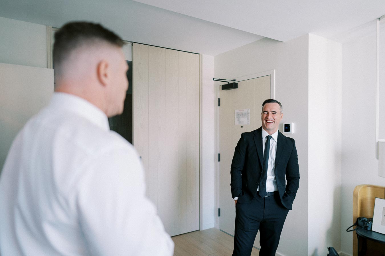 Two men dressed in formal attire, standing in a well-lit room, one smiling and engaging the other in conversation.