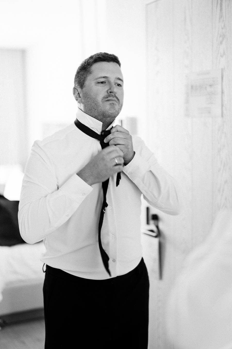 Man in a white dress shirt adjusting a black necktie in front of a mirror, preparing for a formal event.