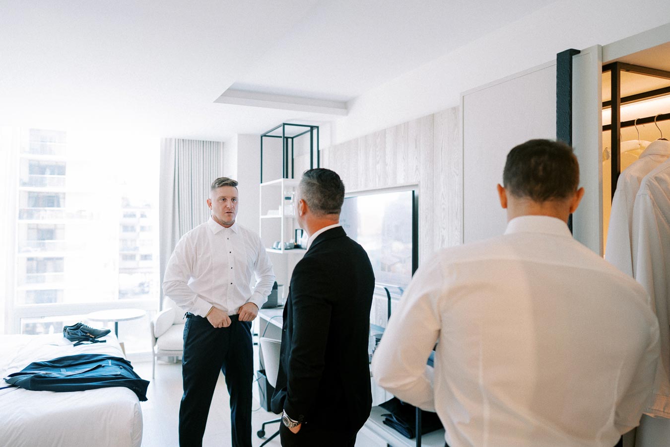 Groom preparing for wedding day in hotel room with two groomsmen in white shirts and suits, discussing plans with window view in background.