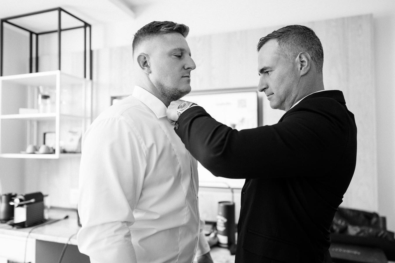 Black and white image of two men, one adjusting the other's tie, in a modern room with shelves and decor, capturing a moment of preparation and focus.
