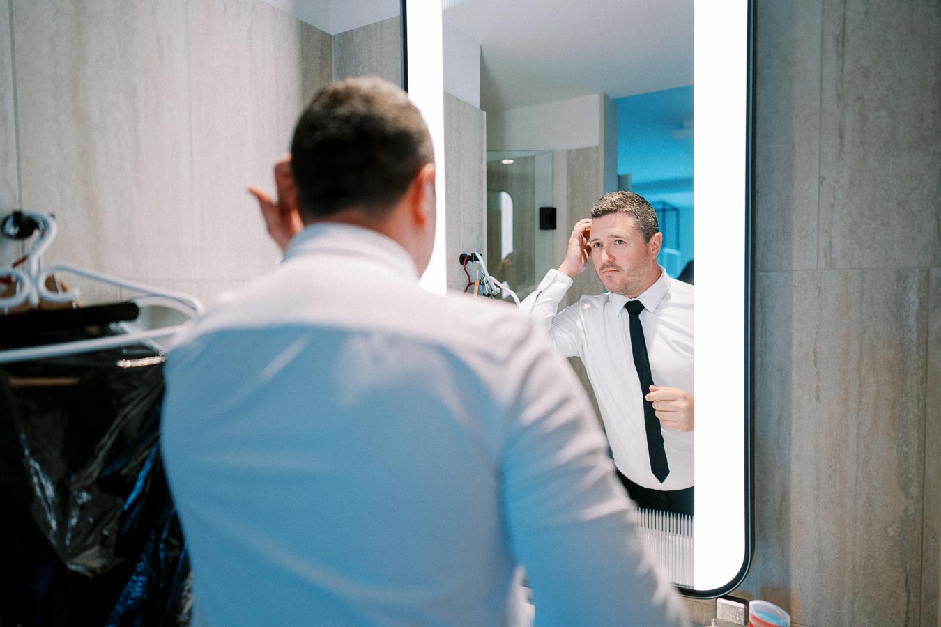 Man adjusting his hair in front of a brightly lit bathroom mirror, dressed in a white shirt and black tie, preparing for a formal event.