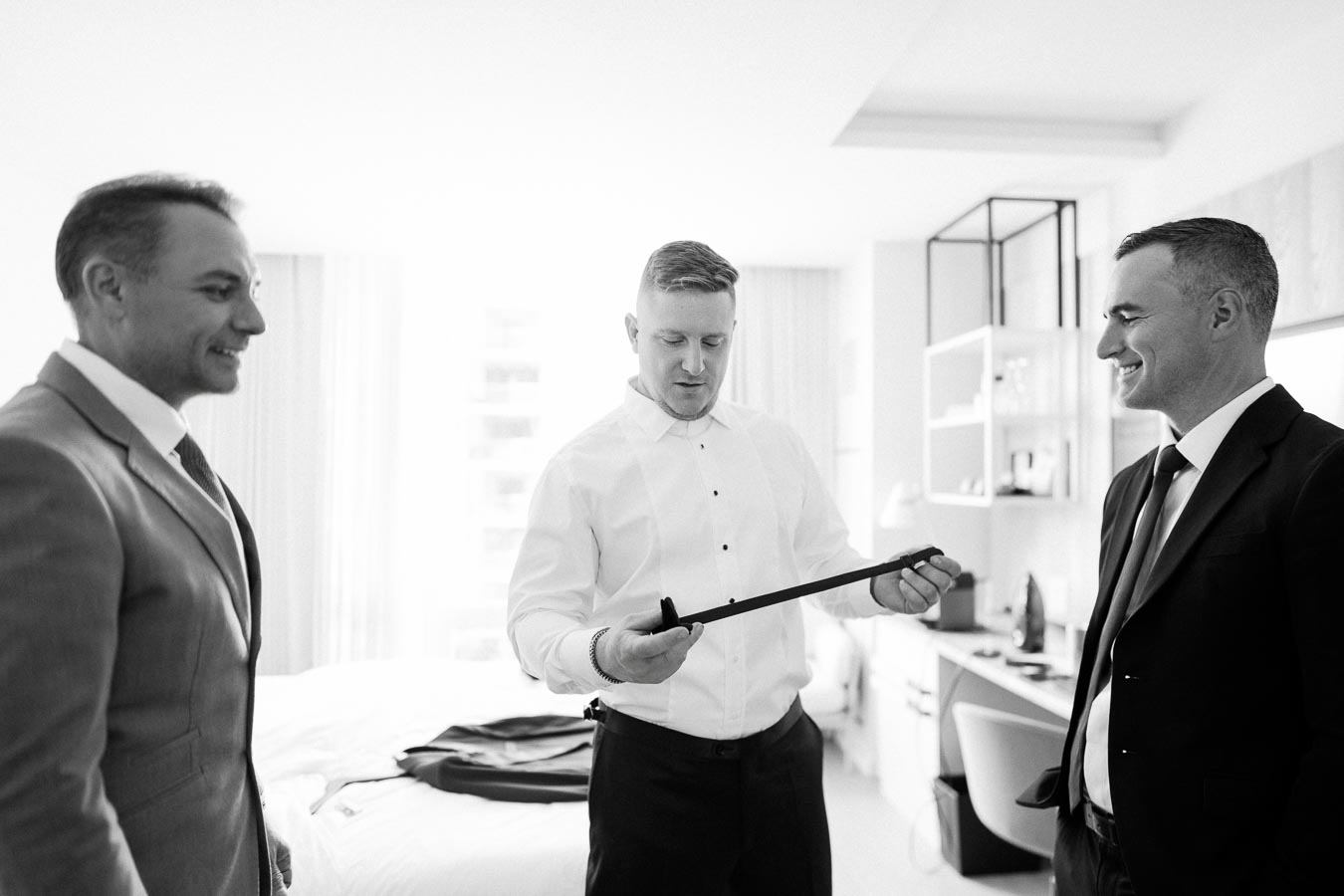 Three men in suits getting ready for a formal event in a bright hotel room, with one man holding a tie.