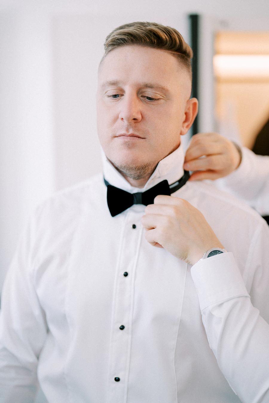 A man in a white dress shirt getting ready, adjusting his black bow tie, wearing a wristwatch, preparing for a formal event.