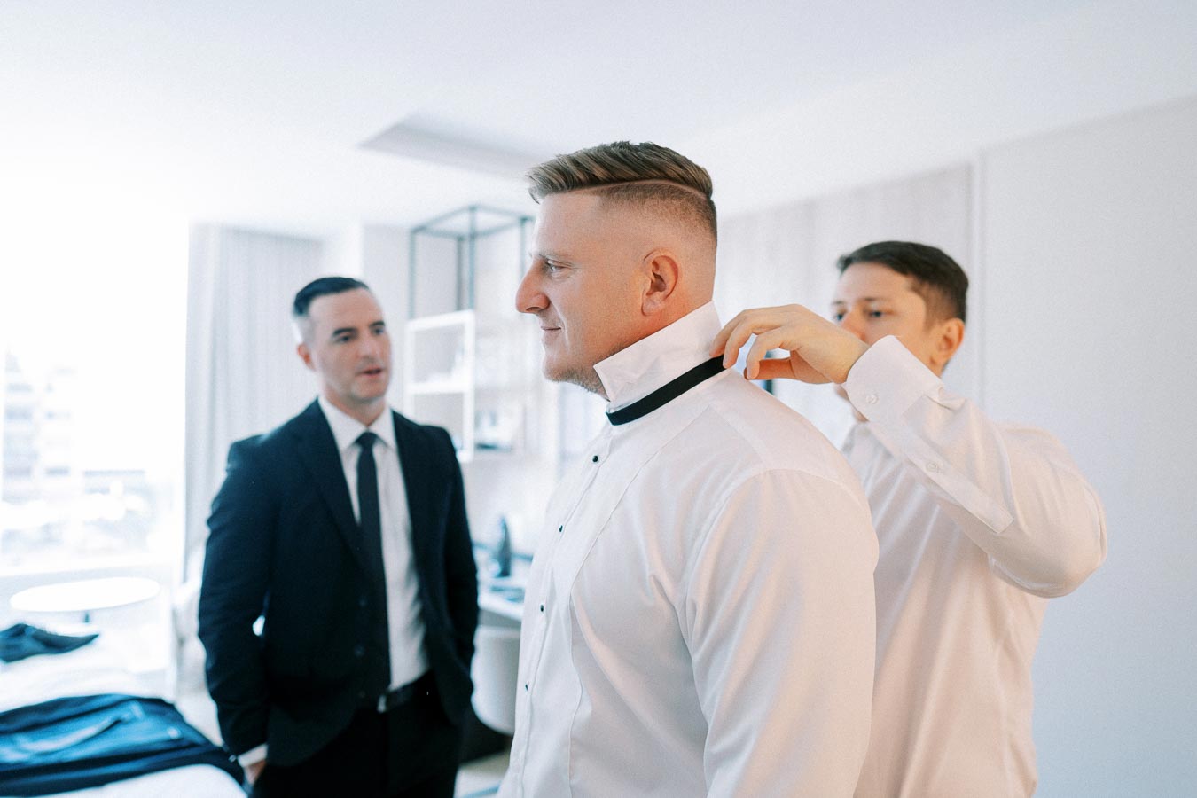 Three men standing in a bright room, two wearing formal suits, one adjusting the bow tie of the third man in a white dress shirt, preparing for a formal event or wedding.