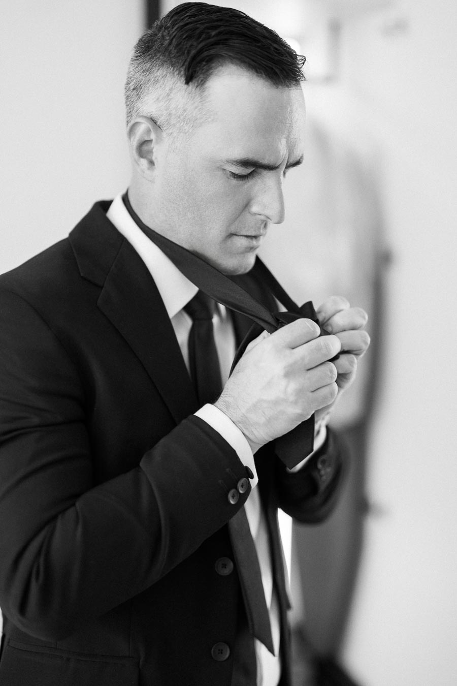 Black and white photo of a man in a suit adjusting his tie, highlighting professional attire and preparation.