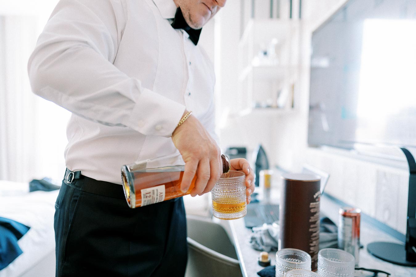 Man in a tuxedo pouring whiskey into a glass, preparing a drink in a modern room setting.