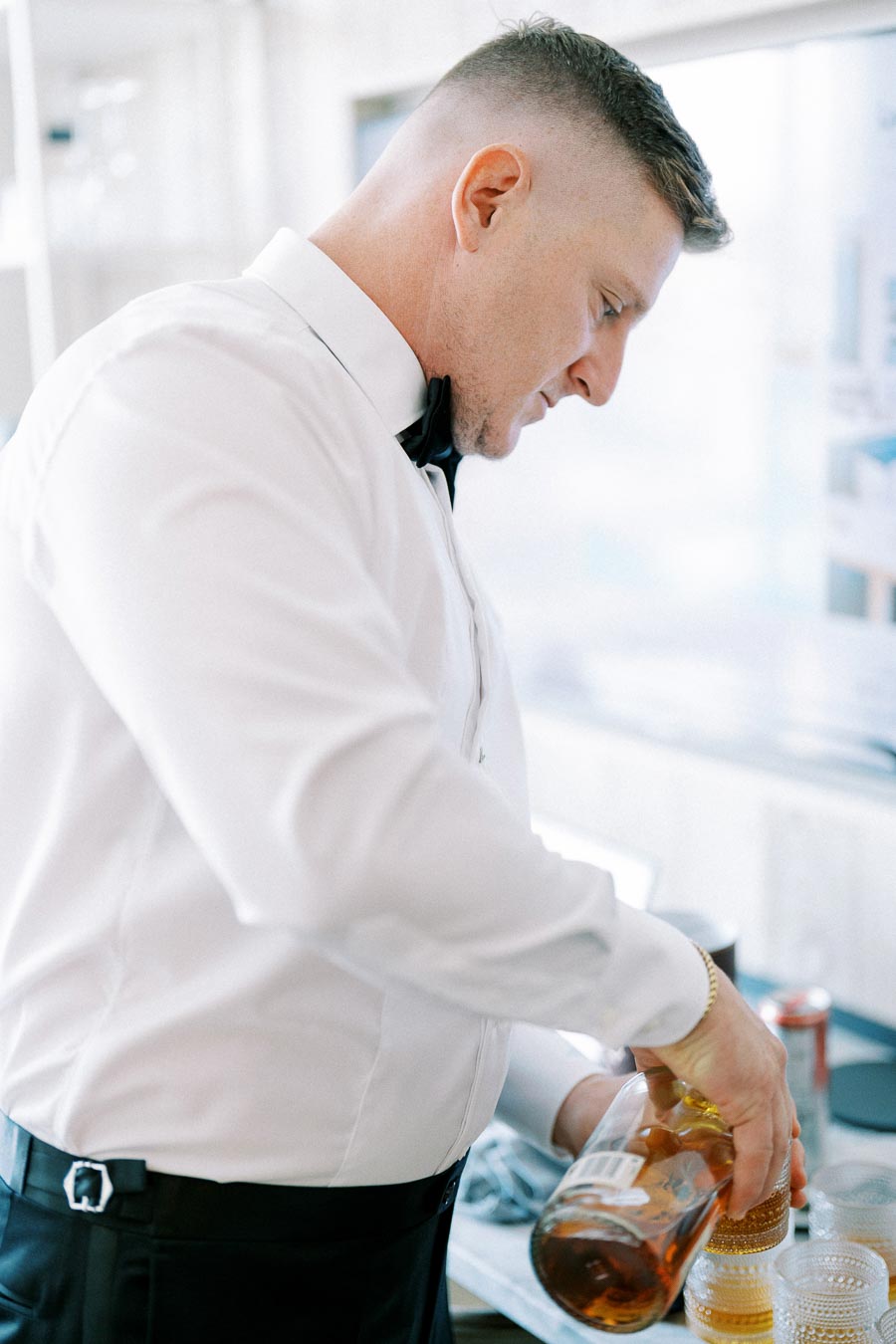 Man in a white dress shirt and bow tie pouring a drink into an elegant glass, likely preparing for a formal event or celebration.